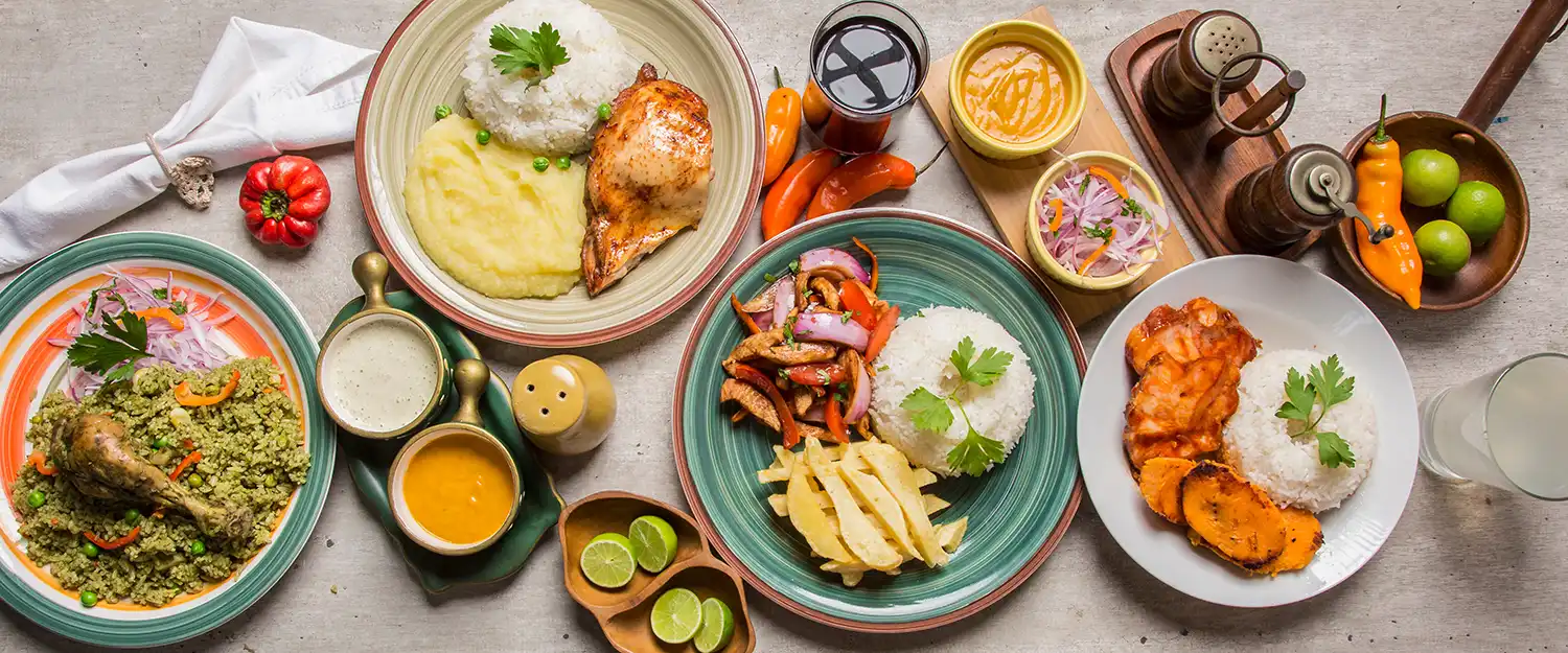 Draufsicht auf peruanische Gerichte: Arroz con Pato, Lomo Saltado mit Pommes, gegrillter Fisch, Ají-Amarillo-Saucen, rote Zwiebeln und Limetten.